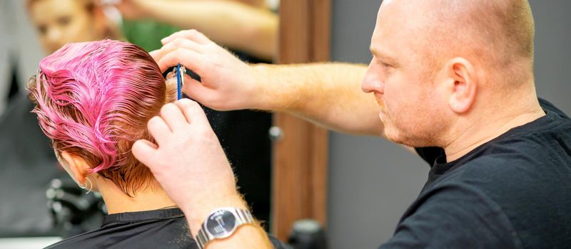 Haircut of dyed short pink wet hair of young caucasian woman by a male hairdresser in a barbershop