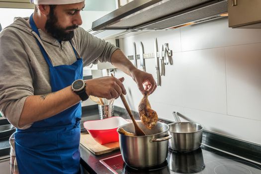 bearded man and apron cooking chicken in the pot with a wooden spoon, in the home kitchen