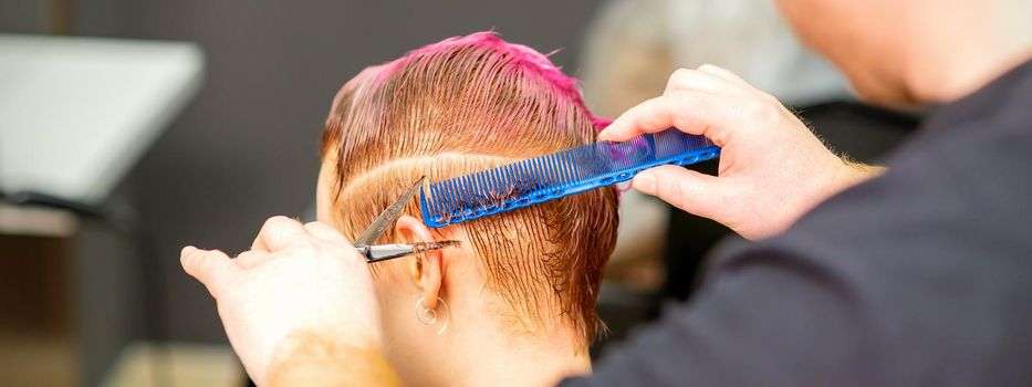 Haircut of dyed short pink wet hair of young caucasian woman by a male hairdresser in a barbershop