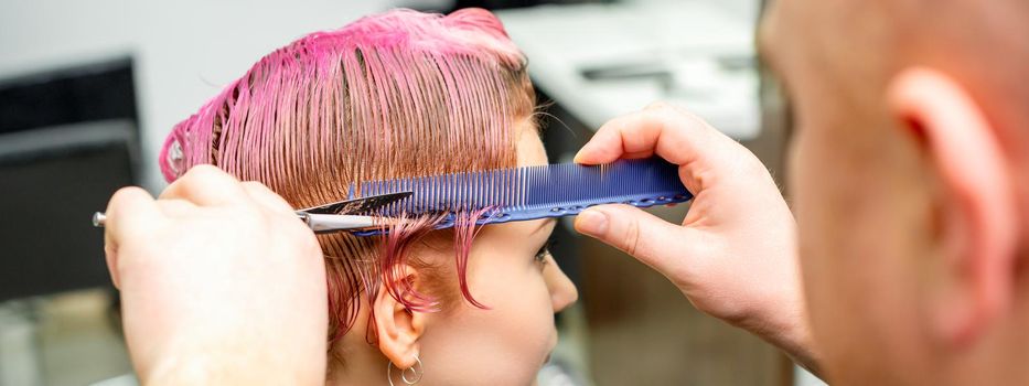 Haircut of dyed short pink wet hair of young caucasian woman by a male hairdresser in a barbershop