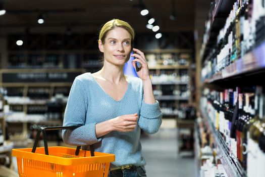 A young beautiful blonde woman stands at the alcohol department with a basket for goods, talks on the phone, consults, asks which alcoholic drink to buy. He looks at the assortment, chooses, smiles.