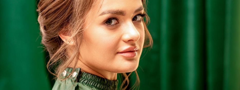 The fashionable young woman. Portrait of the beautiful female model with long hair and makeup with closed eyes. Beauty young woman with a brown curly hairstyle on the background of a green curtain