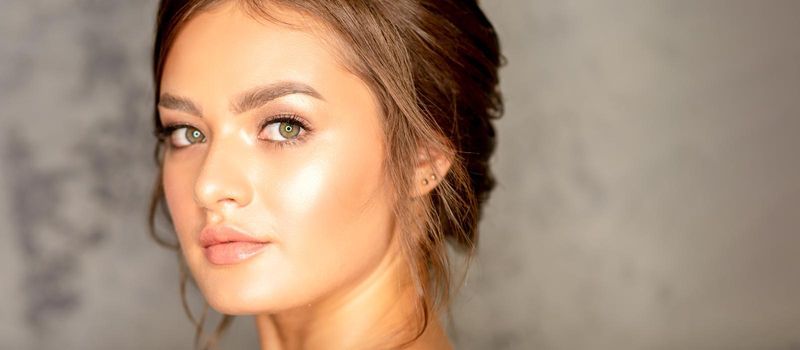 The fashionable young woman. Portrait of the beautiful female model with long hair and makeup. Beauty young caucasian woman with a brown curly hairstyle on the background of a gray wall