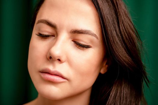 The fashionable young woman. Portrait of the beautiful female model with long hair and makeup with closed eyes. Beauty young woman with a brown curly hairstyle on the background of a green curtain