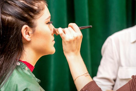 The face of the young woman receives powder on her nose with a brush with the hand of a makeup artist