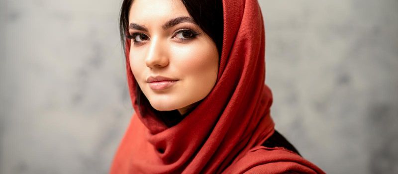 The fashionable young woman. Portrait of the beautiful female model with long hair and makeup in a red scarf. Beauty young caucasian woman on the background of a gray wall