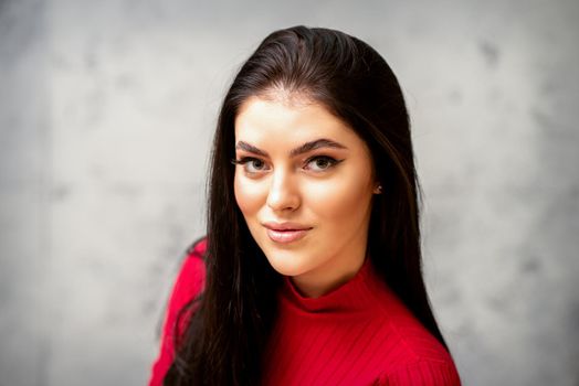 The fashionable young woman. Portrait of the beautiful female model with long hair and makeup. Beauty young caucasian woman with a black hairstyle on the background of a gray wall