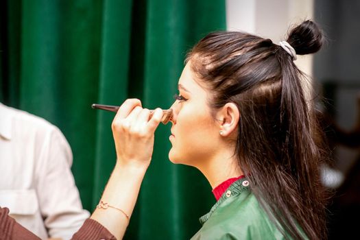 The face of the young woman receives powder on her nose with a brush with the hand of a makeup artist