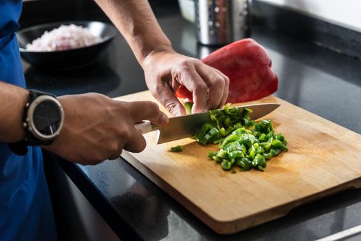 close-up of male hands chopping green and red pepper on a wooden board with a chef's knife, he has other vegetables needed for his recipe in the background