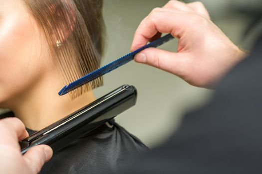 Hairstylist is straightening short hair of young brunette woman with a flat iron in a hairdresser salon, close up