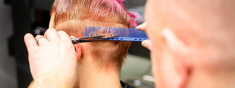 Haircut of dyed short pink wet hair of young caucasian woman by a male hairdresser in a barbershop