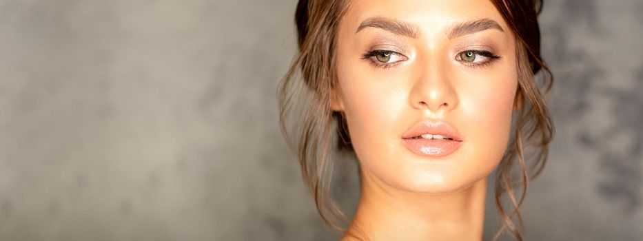 The fashionable young woman. Portrait of the beautiful female model with long hair and makeup. Beauty young caucasian woman with a brown curly hairstyle on the background of a gray wall