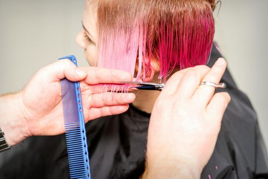 Haircut of dyed short pink wet hair of young caucasian woman by a male hairdresser in a barbershop