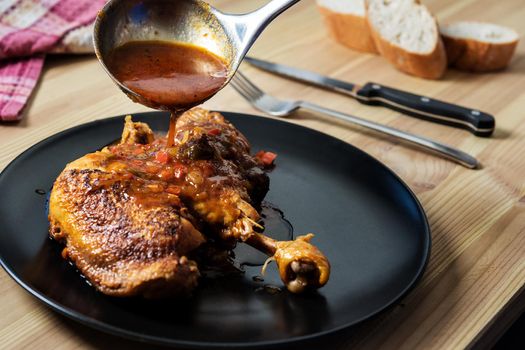 ladle pouring vegetable garnish in sauce over the chicken on the plate. On the wooden table there are cutlery, bread and a vintage kitchen rag