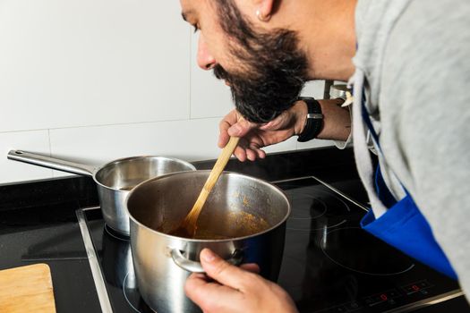 young amateur cook with a beard, smells the food he is preparing in the pot while moving it with the wooden spoon