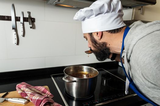 bearded man, chef hat and apron tastes what he is cooking in the home kitchen