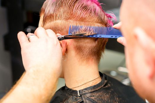 Haircut of dyed short pink wet hair of young caucasian woman by a male hairdresser in a barbershop