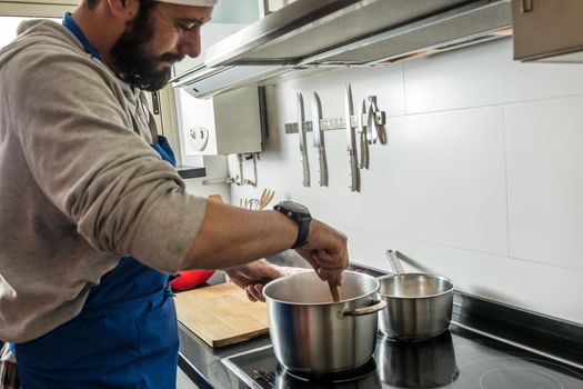 man with beard and apron stirring the pot with a wooden spoon, in the home kitchen