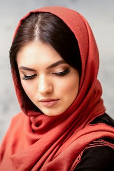The fashionable young woman. Portrait of the beautiful female model with long hair and makeup with eyelash extensions in a red scarf. Beauty young caucasian woman on the background of a gray wall