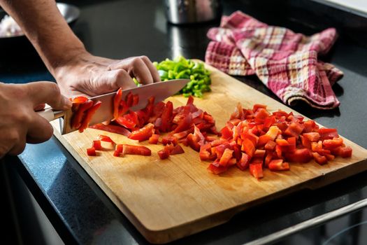 chef's hands chopping red pepper on a wooden cutting board, in the background you see chopped green pepper and a kitchen rag