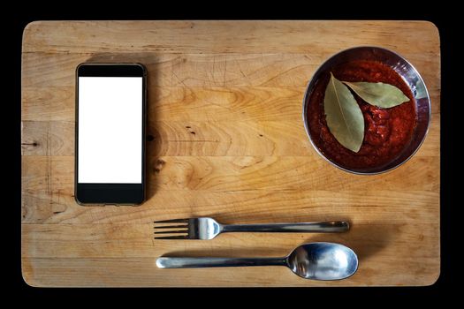 old wooden board with tomato sauce and laurel leaves in a bowl, with cutlery and a smart-phone. Flat lay, top view with copy space