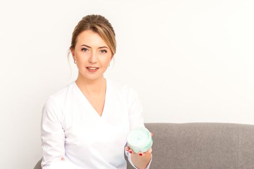 Close-up portrait of young smiling female caucasian healthcare worker standing with jar of cream beauty product on white background.
