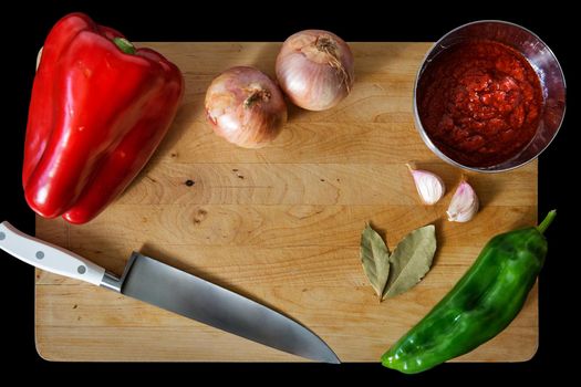wooden cutting board with peppers, onions, garlic, bay leaves, tomato sauce in a bowl and a chef's knife. Top view with copy space