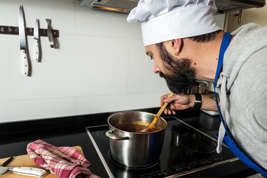 man with beard, chef hat and apron smells what he is cooking in the home kitchen