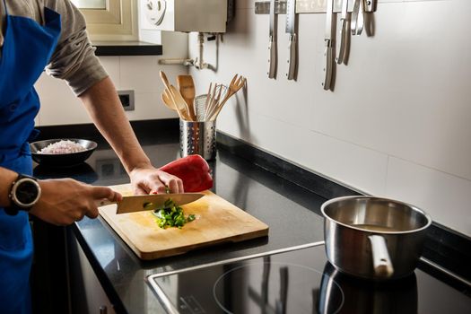 male hands chopping green and red pepper on a wooden board with a chef's knife. He has vegetables and kitchen utensils needed for his recipe