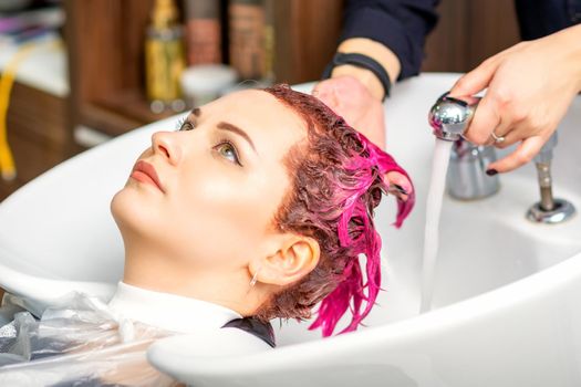 Washing dyed female hair. A young caucasian woman having her hair washed in a beauty salon. Professional hairdresser washes pink color paint off of a customer's hair