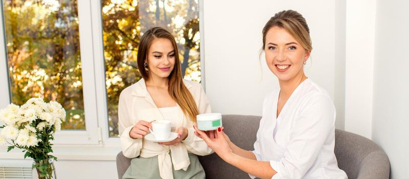 Beautician offering product for the young woman holding a white plastic jar with a cream sitting on the sofa