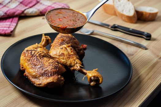 ladle serving vegetable garnish in sauce over the chicken on the plate. On the wooden table there are cutlery, bread and a vintage napkin