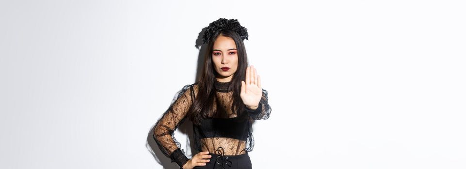 Image of serious asian woman in halloween costume of witch, showing stop gesture, forbid or prohibit something with displeased confident face, standing white background.