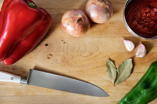 closeup of a wooden cutting board with peppers, onions, garlic, bay leaves, tomato sauce in a bowl and a chef's knife. Top view with copy space
