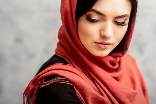 The fashionable young woman. Portrait of the beautiful female model with long hair and makeup with eyelash extensions in a red scarf. Beauty young caucasian woman on the background of a gray wall