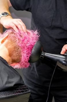 Drying short pink bob hairstyle of a young caucasian woman with a black hair dryer with the brush by hands of a male hairdresser in a hair salon, close up