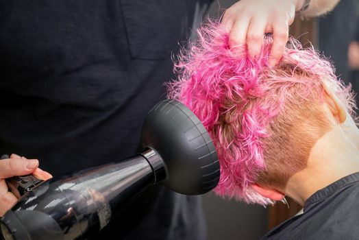 Drying short pink bob hairstyle of a young caucasian woman with a black hair dryer with the brush by hands of a male hairdresser in a hair salon, close up