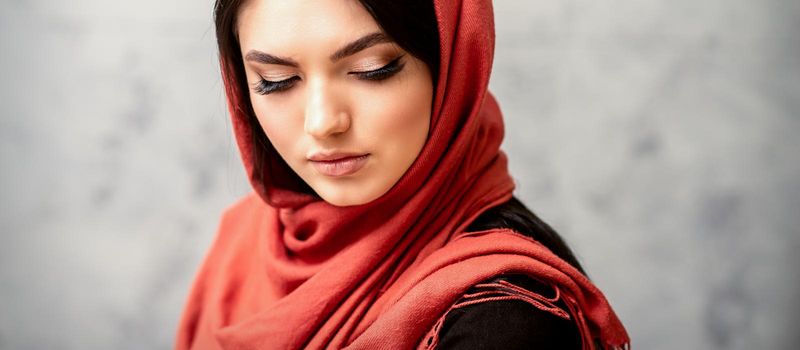 The fashionable young woman. Portrait of the beautiful female model with long hair and makeup with eyelash extensions in a red scarf. Beauty young caucasian woman on the background of a gray wall