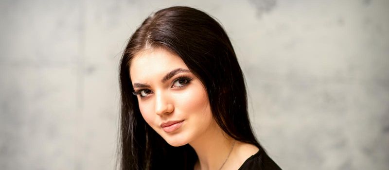 The fashionable young woman. Portrait of the beautiful female model with long hair and makeup. Beauty young caucasian woman with a black hairstyle on the background of a gray wall