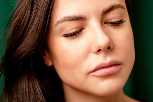 The fashionable young woman. Portrait of the beautiful female model with long hair and makeup with closed eyes. Beauty young woman with a brown curly hairstyle on the background of a green curtain