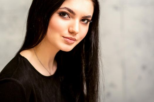 The fashionable young woman. Portrait of the beautiful female model with long hair and makeup. Beauty young caucasian woman with a black hairstyle on the background of a gray wall