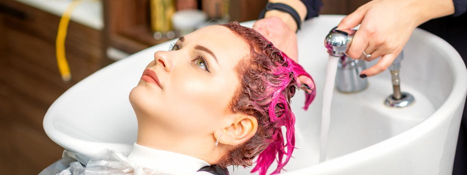 Washing dyed female hair. A young caucasian woman having her hair washed in a beauty salon. Professional hairdresser washes pink color paint off of a customer's hair