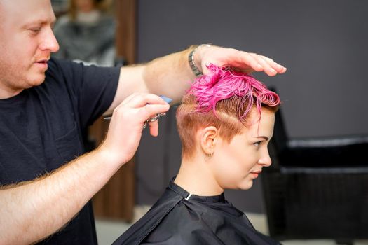 Haircut of dyed short pink wet hair of young caucasian woman by a male hairdresser in a barbershop