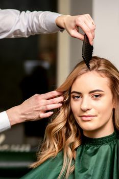 Hairdresser combing long hair of young caucasian woman looking at the camera and smiling in beauty salon