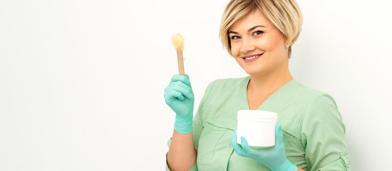 The master of sugar hair removal holds liquid yellow sugar paste, wax for depilation with the jar on a wooden stick on a white background