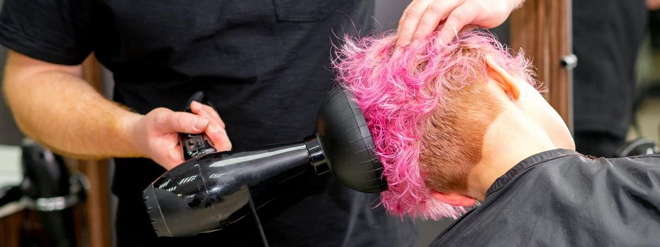 Drying short pink bob hairstyle of a young caucasian woman with a black hair dryer with the brush by hands of a male hairdresser in a hair salon, close up