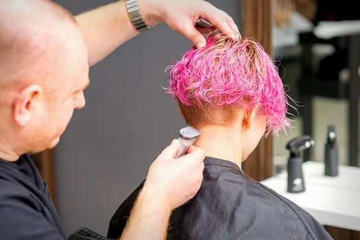 Male hairdresser shaves neck of a young caucasian woman with a short pink hairstyle by electric shaver in a hairdresser salon, close up