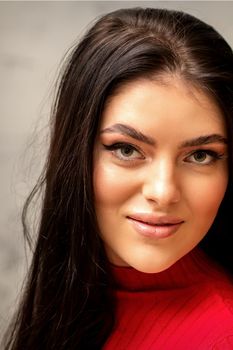 The fashionable young woman. Portrait of the beautiful female model with long hair and makeup. Beauty young caucasian woman with a brown curly hairstyle on the background of a gray wall