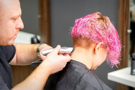 Male hairdresser shaves neck of a young caucasian woman with a short pink hairstyle by electric shaver in a hairdresser salon, close up