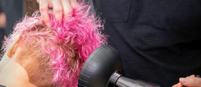 Drying short pink bob hairstyle of a young caucasian woman with a black hair dryer with the brush by hands of a male hairdresser in a hair salon, close up